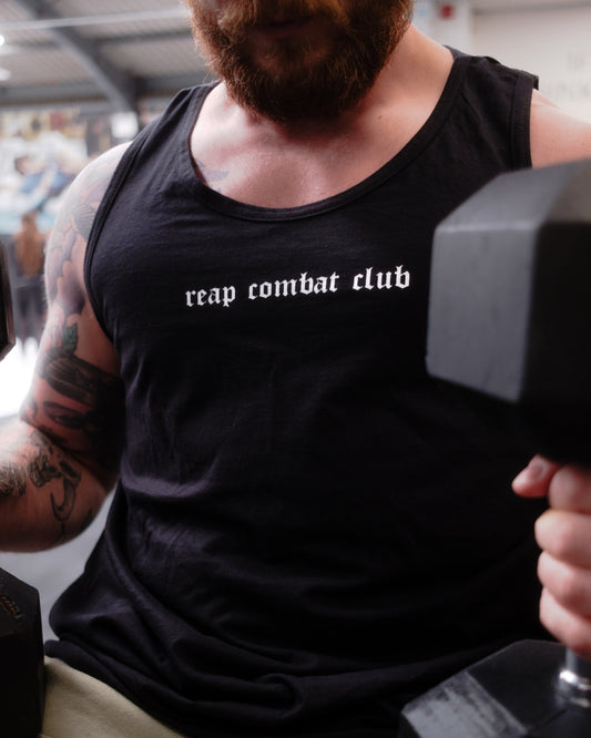 Muscular man lifting dumbbells in a gym wearing a black tank top with ‘reap combat club’ logo, showcasing fitness and combat sports lifestyle apparel