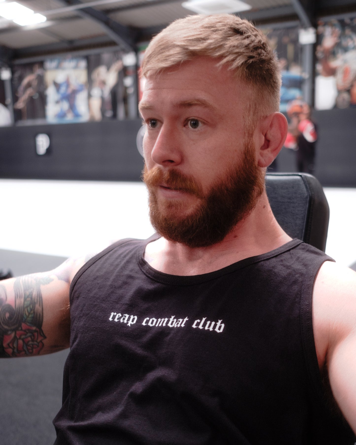 Muscular man lifting dumbbells in a gym wearing a black tank top with ‘reap combat club’ logo, showcasing fitness and combat sports lifestyle apparel