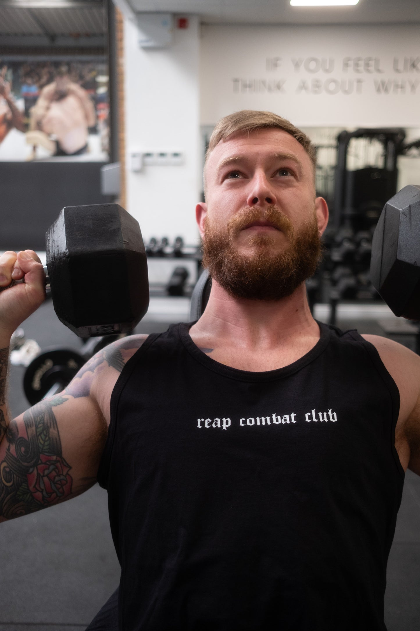 Muscular man lifting dumbbells in a gym wearing a black tank top with ‘reap combat club’ logo, showcasing fitness and combat sports lifestyle apparel