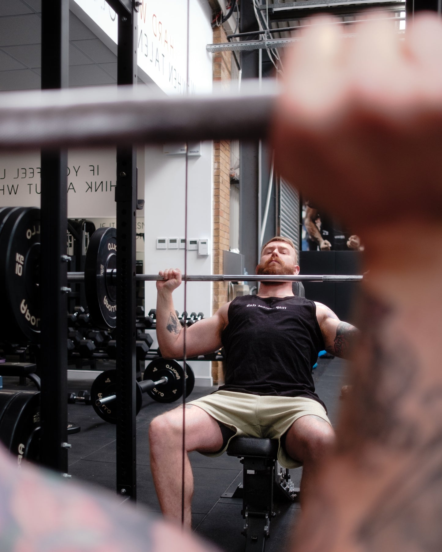 Muscular man lifting dumbbells in a gym wearing a black tank top with ‘reap combat club’ logo, showcasing fitness and combat sports lifestyle apparel