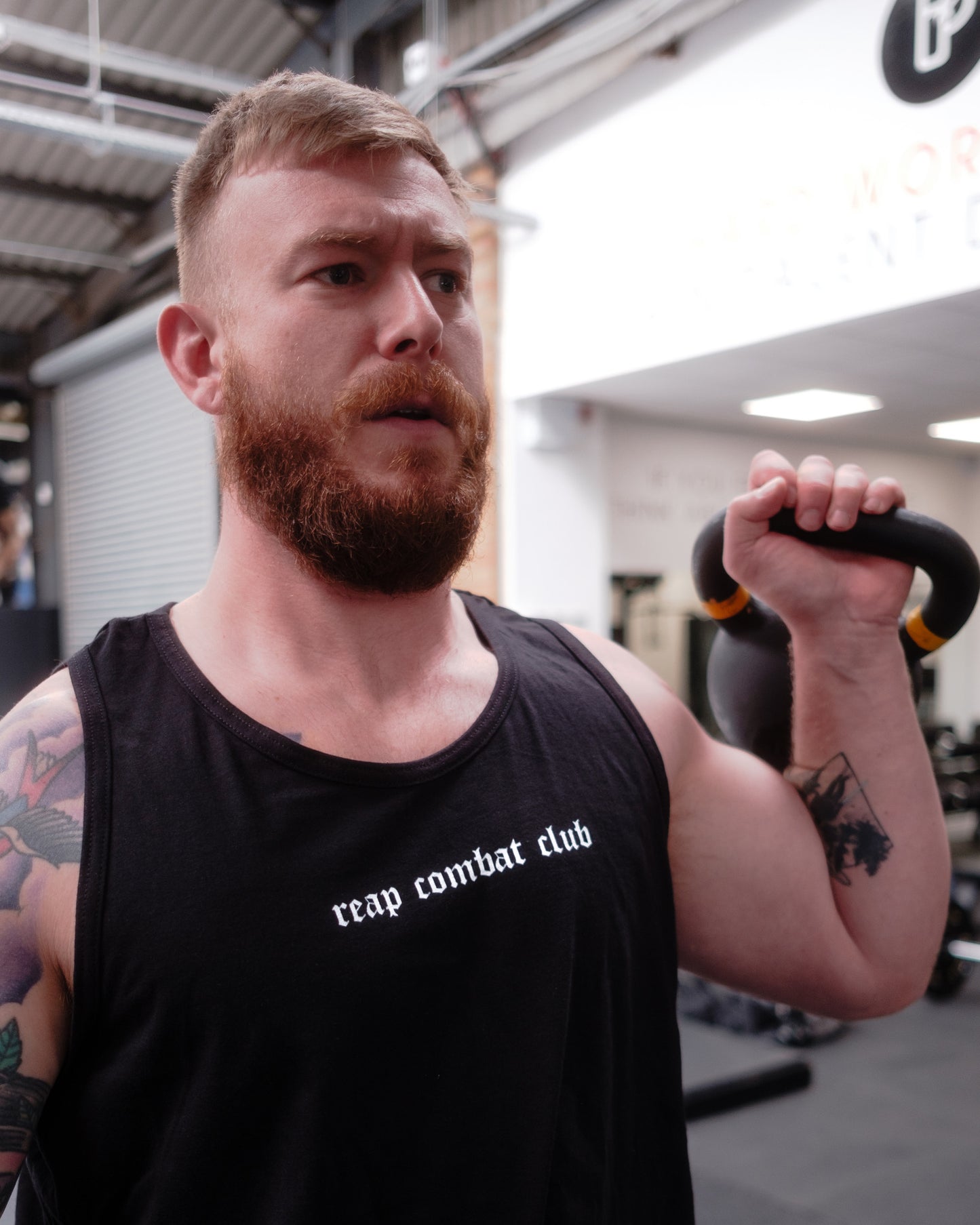 Muscular man lifting dumbbells in a gym wearing a black tank top with ‘reap combat club’ logo, showcasing fitness and combat sports lifestyle apparel