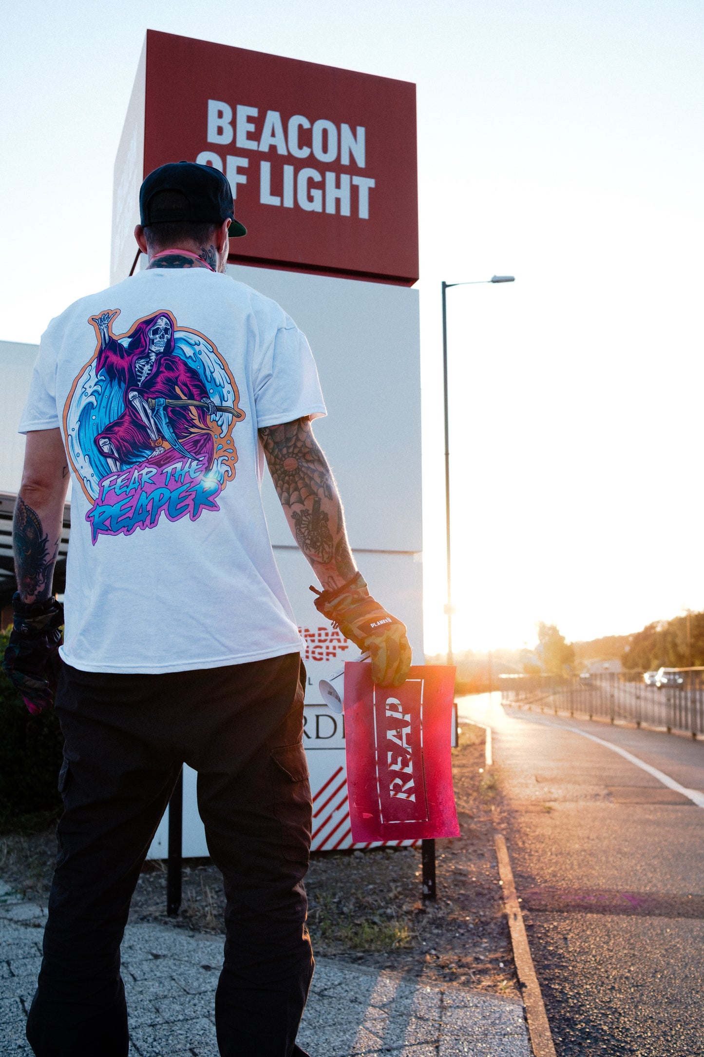 Person wearing a white t-shirt with colorful graphics standing in front of a large sign reading 'Beacon of Light'.