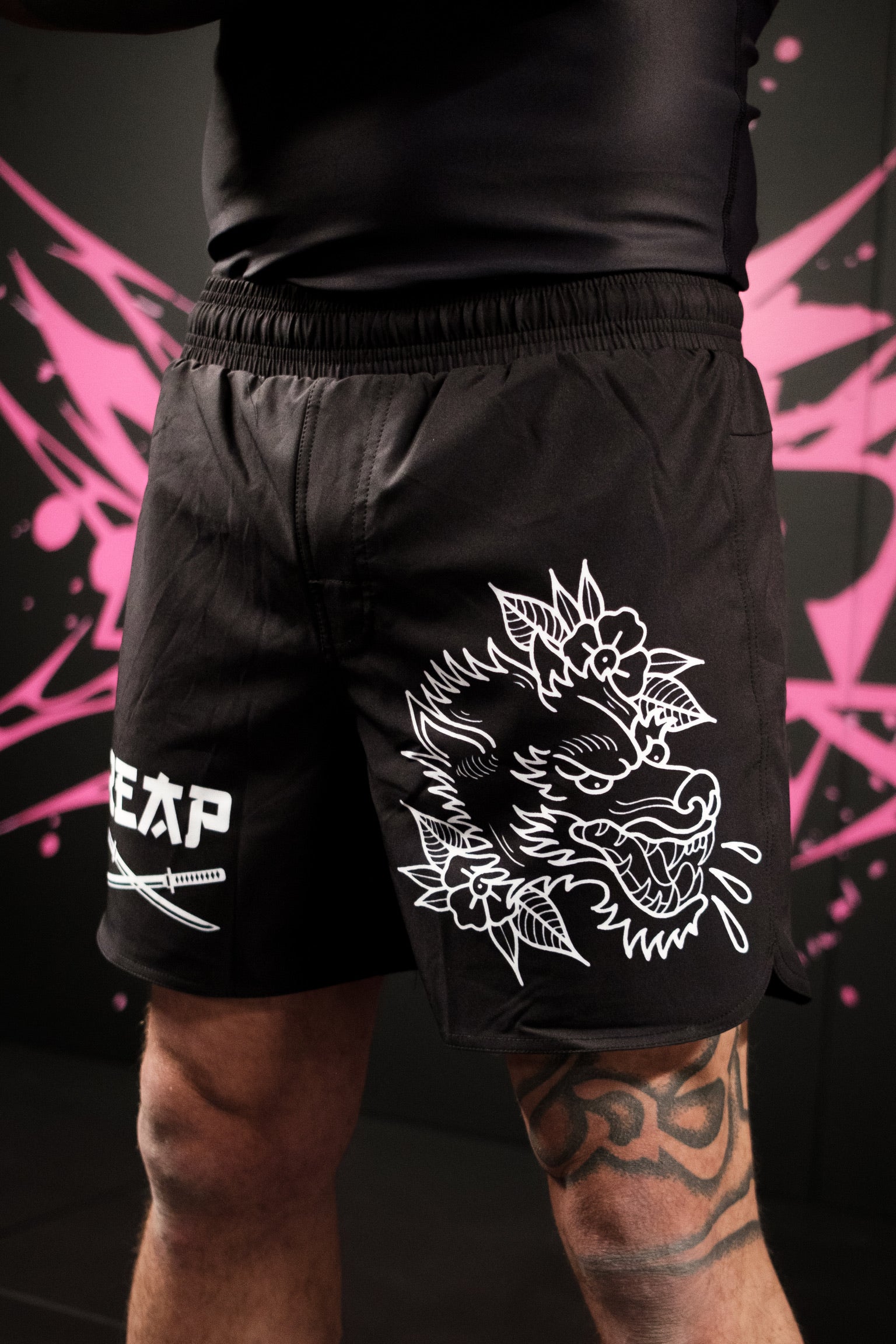 Black shorts with white dragon design and brand logo on a dark background with pink abstract patterns.