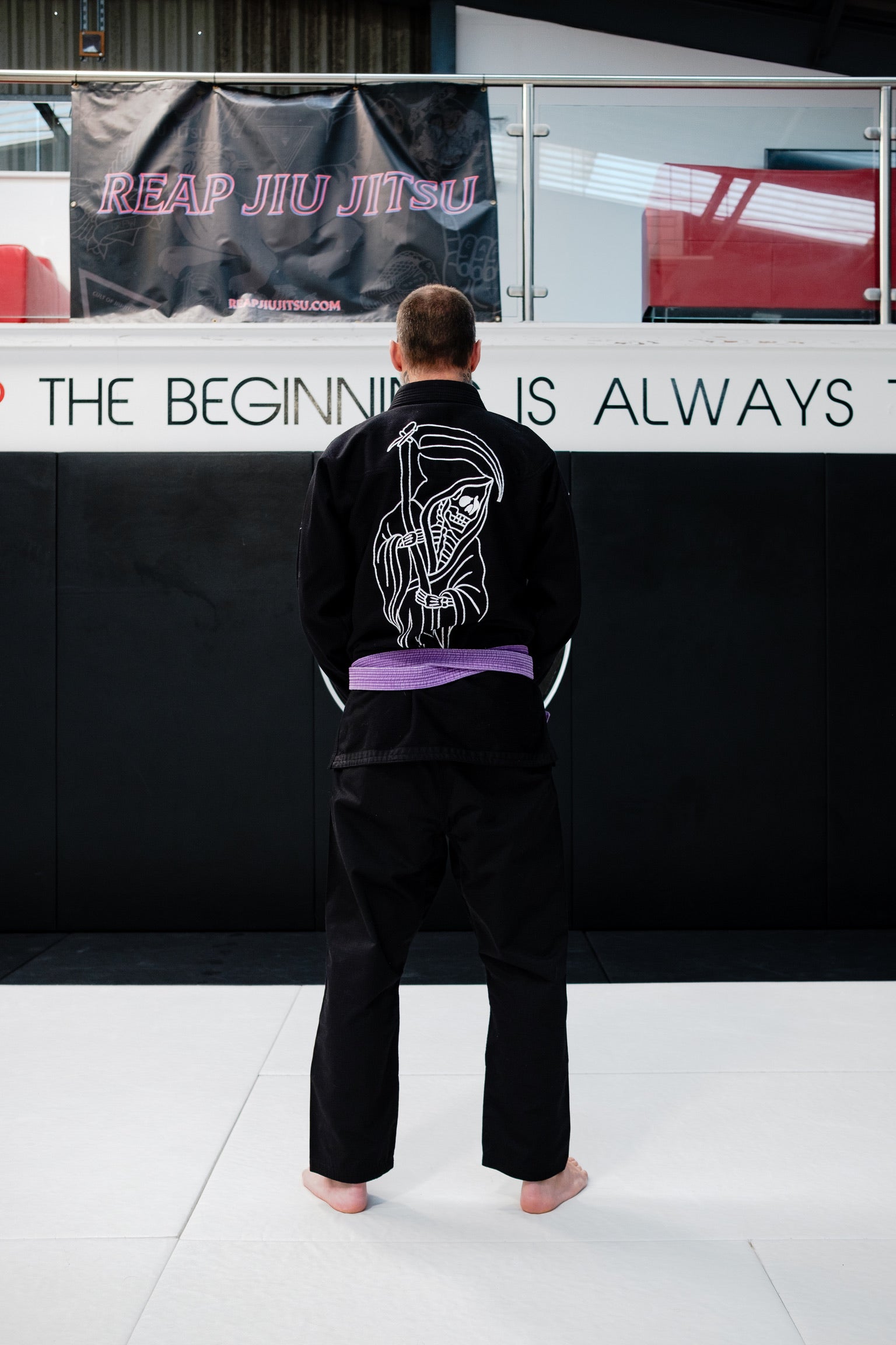 Person wearing a black martial arts uniform with a logo on the back, standing in front of a Reap Jiu Jitsu banner.
