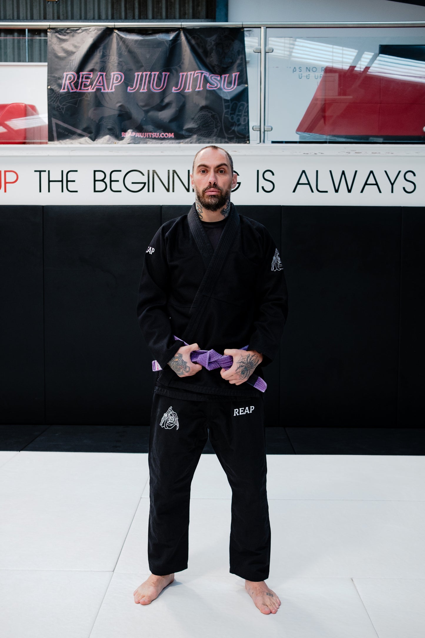 Person in black martial arts uniform with purple belt standing in front of a Reap Jiu Jitsu banner.