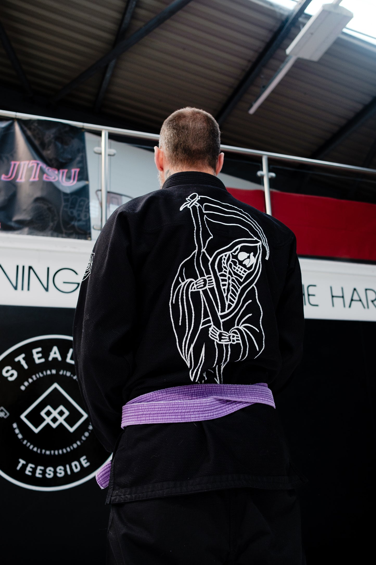 Person wearing a black martial arts gi with a white design on the back, standing in front of a banner.